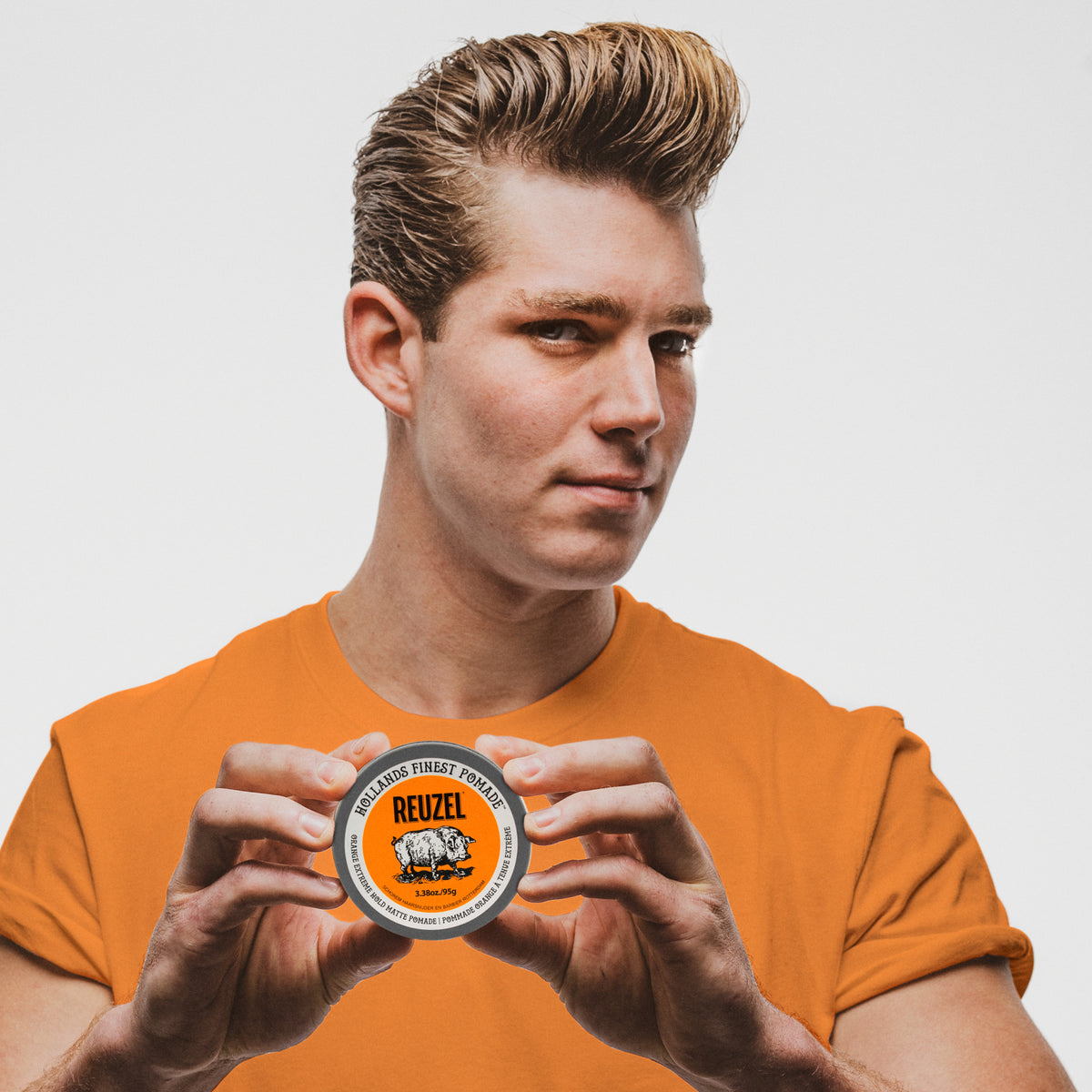Male model with styled hair wearing an orange shirt. Reuzel Orange Extreme Hold matte pomade, showing a container of an orange pomade container for matte finish for strong hold styling.