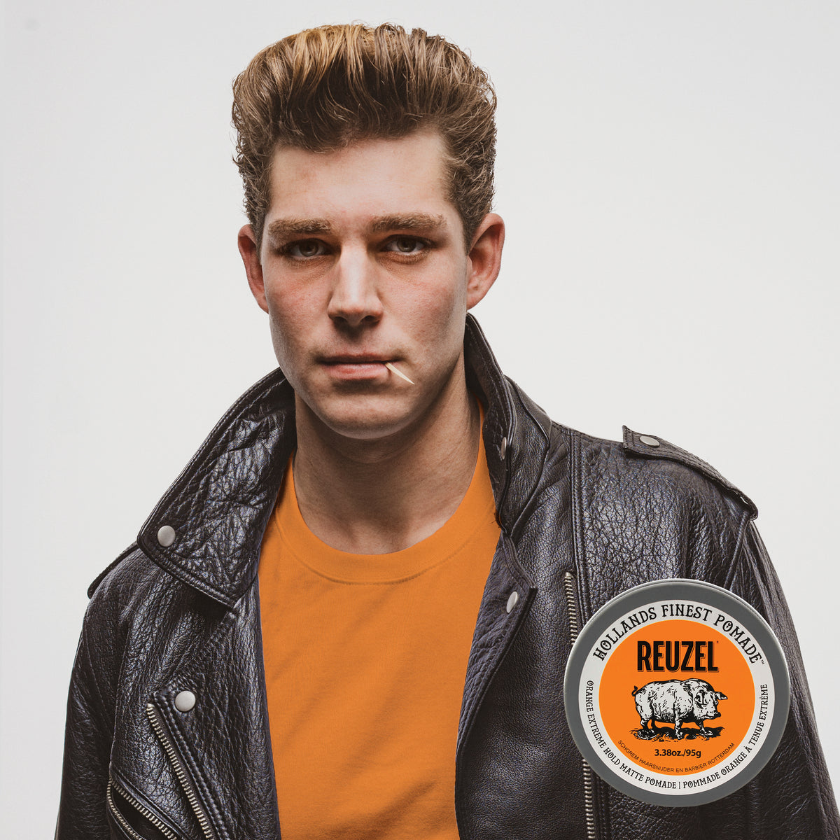 A male model wearing a leather jacked, with hair styled with Orange REUZEL Extreme Hold Matte Pomade jar with orange label, showcasing the product's bold packaging and orange color. 
