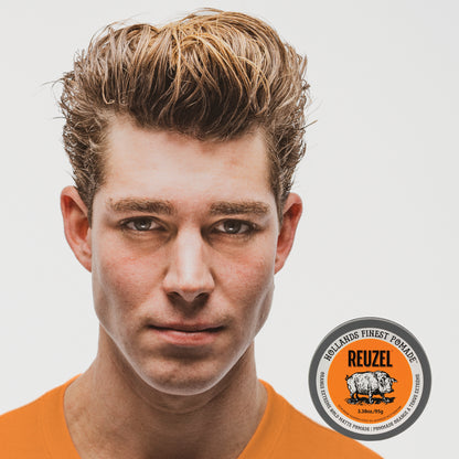 A close-up portrait of a man with styled light brown hair wearing an orange shirt, with a Reuzel Orange Extreme Hold Matte Pomade tin in the lower right corner on a light gray background.