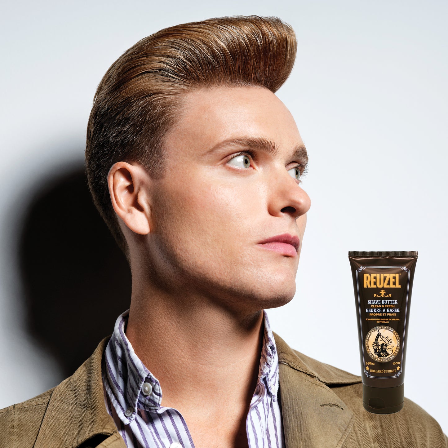 Man with styled hair next to a bottle of Reuzel Shave Butter on a gray background