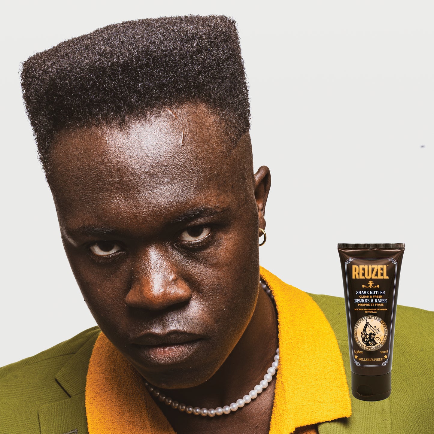 Man with styled hair wearing a green jacket with a yellow collar, next to a Reuzel Shave Butter tube.