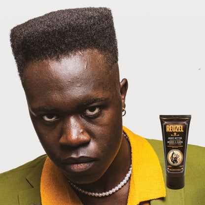 Man with styled hair wearing a green jacket with a yellow collar, next to a Reuzel Shave Butter tube.