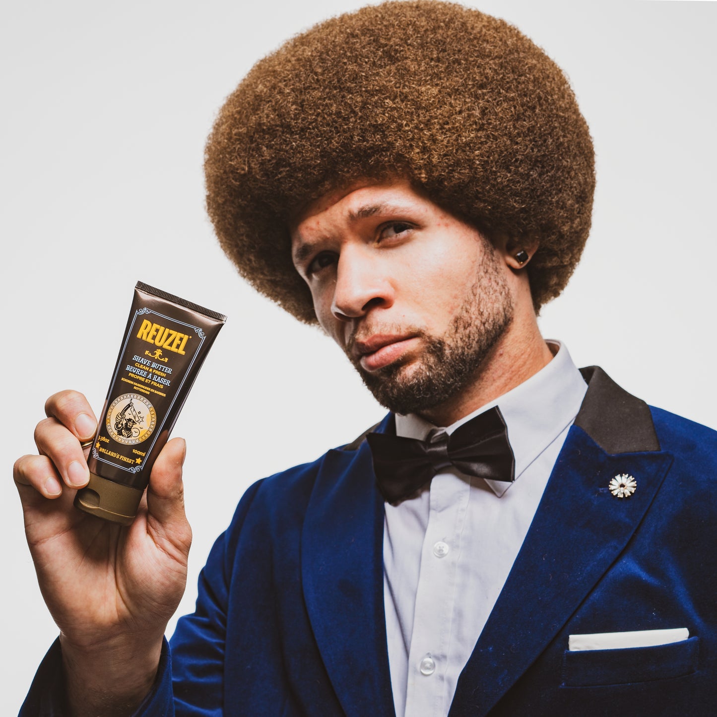 Man in a blue suit holding a Reuzel Shave Butter tube against a plain background